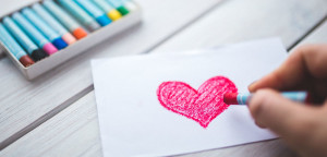 someone coloring a pink heart on a piece of paper using pastels. box of other colors in the distance