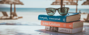 books stacked on a table, sunglasses on top, and a beach in the background out of focus