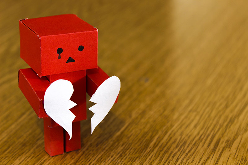 Red, tiny robot made out of paper, a black tear from his right eye, holding a paper heart that's been torn in half