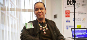 Black woman sitting in a wheelchair wearing a brown outfit and a gold necklace. A laptop with live captions is behind her