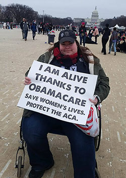 woman sitting in a wheelchair holding a sign that says I am alive thanks to obamacare, protest the ACA. The Capitol building is behind her, in the distance
