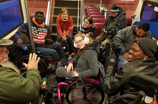 A young woman sits in a wheelchair in winter gear, in a subway car. She is surrounded by five others also bundled up on their way to a protest
