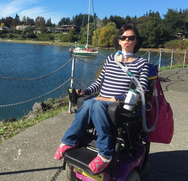 A woman with short hair, a striped shirt, jeans, and pink slippers. She sits in a wheelchair. Behind her is a lake with boats on it.
