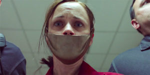 A young woman with her hair messily pulled back and her mouth is covered with a metallic mask. She looks terrified.