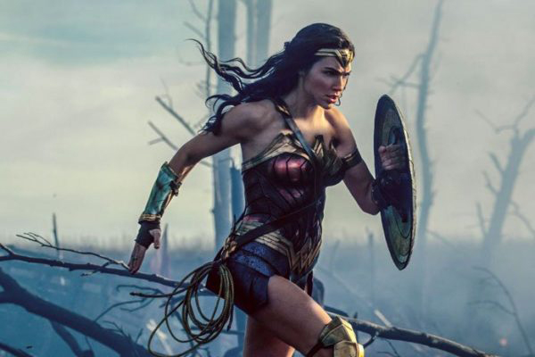 Wonder Woman in her costume, shield up and running, behind her is smoke and burned out trees