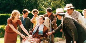 A white man in an old-fashioned wheelchair is outside, abled people surround him, smiling