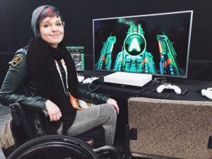 Cherry sitting in her wheelchair, turned to face the camera. On a desk in front of her is a computer, showing a green spaceship